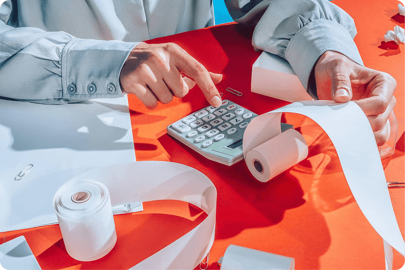 A person's hands using a calculator surrounded by receipt paper rolls on a red desk.